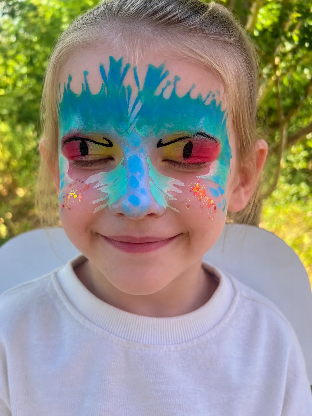 A smiling girl in a turquoise and blue cat mask, with white whiskers across the cheeks, red and yellow eyelids and orange glitter freckles