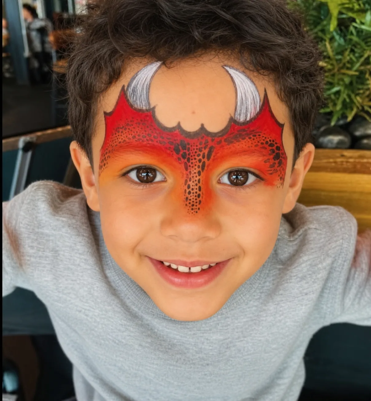 A grinning boy in a red and orange devil mask with white curved horns and fine black scale detail across the brow and nose