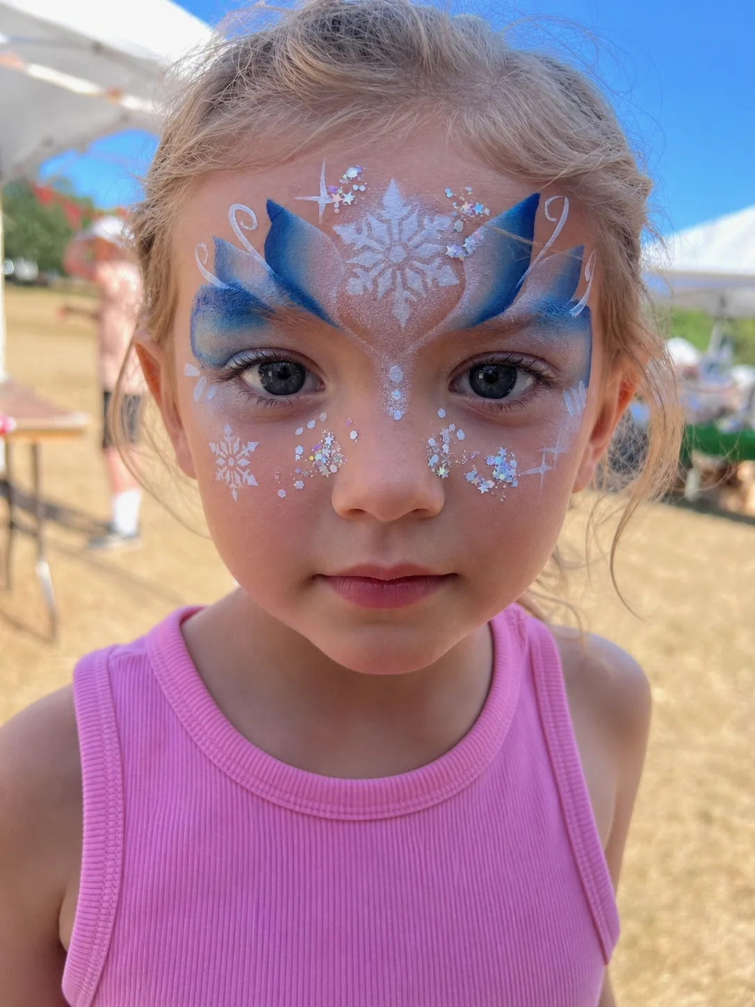 A girl at an outdoor event wearing an icy blue crown design with a white snowflake on the forehead, holographic glitter and small snowflakes on both cheeks