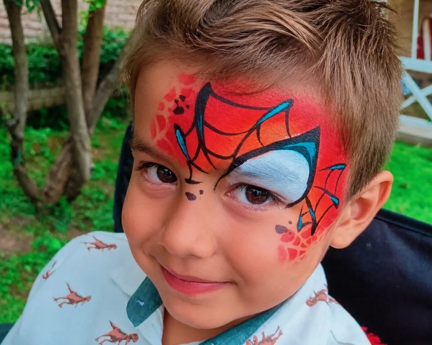 A boy in a garden wearing a red spider-hero mask over one eye, with black web linework and a white and teal eye panel